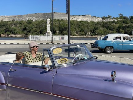 Eine Frau mit Sonnenbrille und Hut fährt in einem lila Oldtimer, während ein Junge neben ihr sitzt und lächelt.