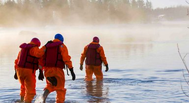 Drei Personen in orangefarbenen Anzügen waten durch einen nebligen Fluss, während die Sonne aufgeht und die Umgebung erhellt.