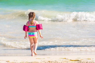 Ein kleines Mädchen mit Zöpfen trägt bunte Schwimmflügel und geht fröhlich zum Wasser am Strand.