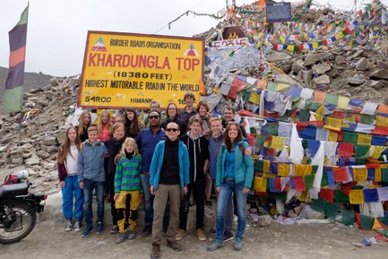 Eine Gruppe von Reisenden posiert fröhlich vor dem Khardungla-Pass-Schild, umgeben von bunten Gebetsfahnen.