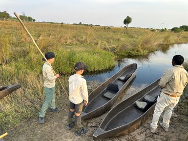 Drei Kinder und ein Erwachsener stehen am Ufer eines ruhigen Gewässers, umgeben von hohem Gras und Booten.