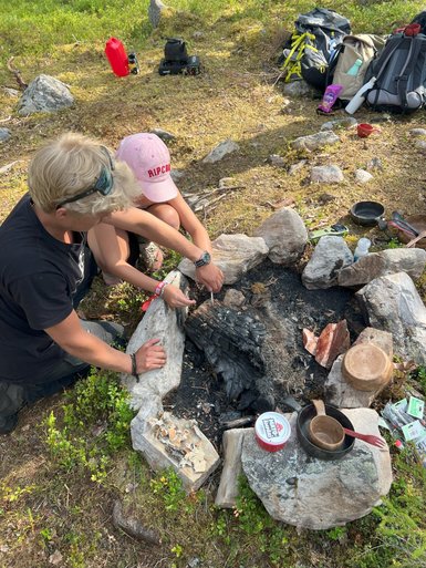 Zwei Personen arbeiten gemeinsam am Lagerfeuer, während sie Holzstücke bearbeiten und sich auf das Kochen vorbereiten.