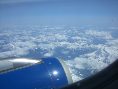 Blick aus dem Flugzeugfenster auf schneebedeckte Berge und Wolken unter einem klaren blauen Himmel.