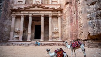 Zwei Kamele mit bunten Sätteln ruhen im Sand vor der beeindruckenden Fassade eines antiken Gebäudes in Petra.