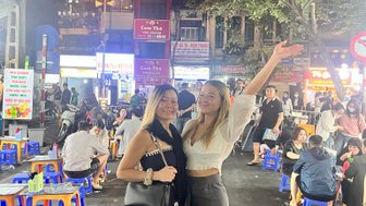 Zwei Frauen posieren fröhlich in einer belebten Straßenmarktszene bei Nacht, umgeben von bunten Stühlen und Lichtern.