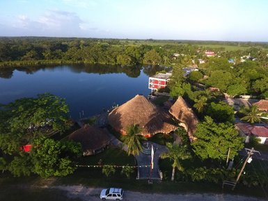 Eine Luftaufnahme zeigt das Lamanai Landings Hotel mit strohgedeckten Hütten am ruhigen Wasser eines Sees.