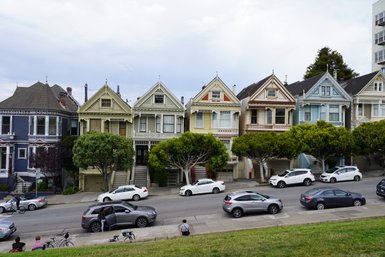 Bunte viktorianische Häuser reihen sich an einer Straße in San Francisco, umgeben von grünen Bäumen und parkenden Autos.