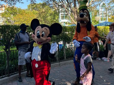 Mickey Mouse und Goofy gehen fröhlich durch einen Park, während ein kleines Mädchen ihnen neugierig folgt.