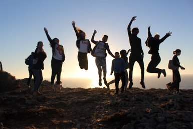 Eine Gruppe von Menschen springt fröhlich auf einem Felsen, während die Sonne im Hintergrund untergeht und den Himmel erleuchtet.