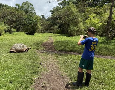 Ein Junge in Gummistiefeln fotografiert eine große Schildkröte auf einem schmalen Pfad in einer grünen Landschaft.