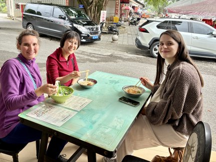 Drei Frauen sitzen an einem Tisch im Freien und genießen ihre Nudelsuppe mit Stäbchen, während sie fröhlich lächeln.