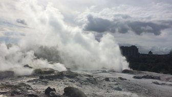 Dampf steigt aus dem geothermalen Gebiet in Rotorua auf, umgeben von grauen Felsen und einer bewölkten Himmel.