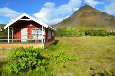Ein rotes Ferienhaus mit einer Veranda steht vor einem majestätischen Berg, umgeben von grünen Wiesen und blühenden Pflanzen.