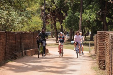 Drei Radfahrer, darunter ein Mann und zwei Mädchen, fahren fröhlich auf einem schmalen Weg zwischen alten Ziegelmauern.