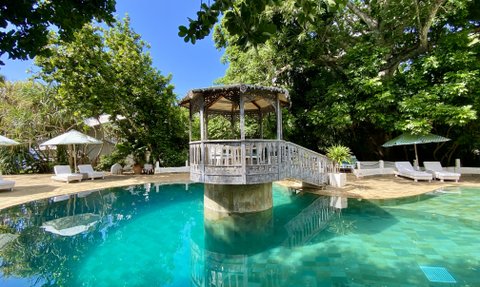 Ein ruhiger Poolbereich mit klarem Wasser, umgeben von üppigem Grün und einem charmanten Pavillon im Hintergrund.