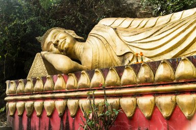 Eine große, goldene Buddha-Statue liegt entspannt auf einer roten Plattform, umgeben von üppigem Grün und kleinen Altären.