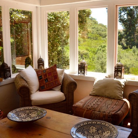 Ein gemütlicher Raum mit Rattanstühlen, bunten Kissen und Blick auf die grüne Landschaft durch große Fenster.