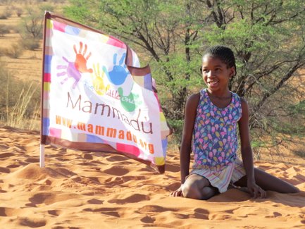 Ein fröhliches Mädchen sitzt im Sand und lächelt, während eine bunte Flagge mit dem Schriftzug 'Mammadu' im Hintergrund weht.