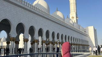 Eine Person in einem roten Gewand steht am Rand eines Wasserspiegels, während die beeindruckende Sheikh Zayed Moschee im Hintergrund sichtbar ist.