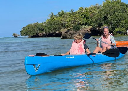 Ein Junge und eine Frau paddeln fröhlich in einem blauen Kajak, umgeben von klarem Wasser und üppigem Grün.