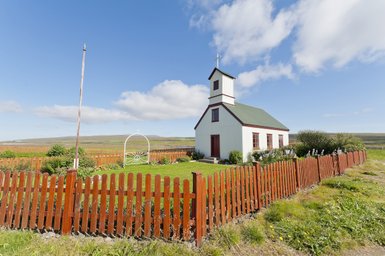 Eine kleine, weiße Kirche mit grünem Dach steht inmitten einer blühenden Wiese, umgeben von einem roten Holzzaun.