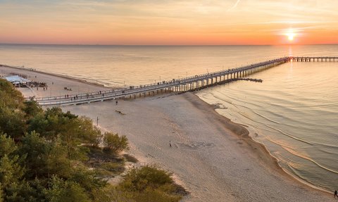 Ein malerischer Sonnenuntergang über dem Meer, während Menschen auf einer langen Seebrücke spazieren gehen.