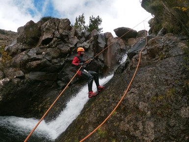 Ein Sportler in einem roten Anzug seilt sich an einer steilen Felswand mit Wasserfall ab, umgeben von Natur.