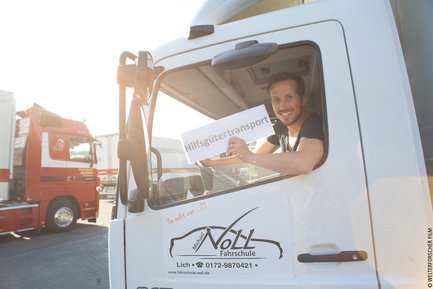 Ein lächelnder Mann sitzt im Fahrerhaus eines Lkw und hält ein Schild mit der Aufschrift 'Hilfsgütertransport' hoch.