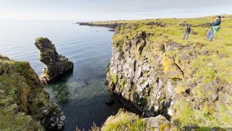 Zwei Wanderer stehen an einer steilen Küste, während das ruhige Wasser und ein markanter Felsen im Hintergrund sichtbar sind.