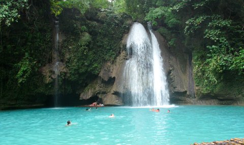 Ein atemberaubender Wasserfall stürzt in ein türkisfarbenes Becken, umgeben von üppigem Grün und fröhlichen Schwimmern.