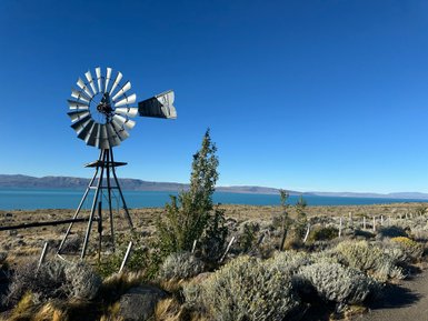 Eine alte Windmühle steht majestätisch vor dem klaren blauen Wasser des Lago Argentino, umgeben von sanften Hügeln.