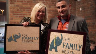 Ein Mann und eine Frau lächeln, während sie zwei gerahmte Bilder mit einem Elefantenmotiv halten, in einem gemütlichen Raum.