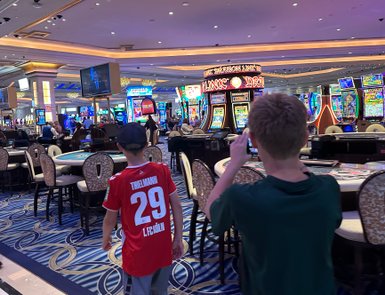 Zwei Kinder erkunden ein Casino, während ein Fernseher im Hintergrund Nachrichten zeigt und Spielautomaten leuchten.