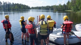 Eine Gruppe von sechs Personen in Schwimmwesten und Helmen steht am Ufer eines ruhigen Gewässers, bereit zum Rafting.