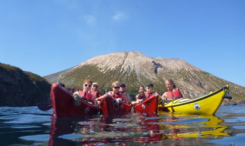 Sechs Kajakfahrer paddeln in ruhigem Wasser, während ein majestätischer Vulkan im Hintergrund thront.