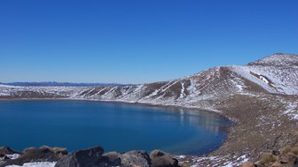 Ein klarer, blauer See umgeben von schneebedeckten Hügeln und einem strahlend blauen Himmel im Tongariro Nationalpark.
