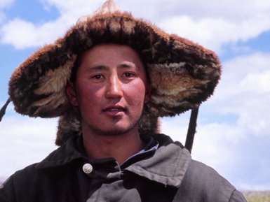 Ein mongolischer Mann trägt einen traditionellen Hut und sitzt mit zwei Kindern in einer grünen Landschaft unter einem blauen Himmel.