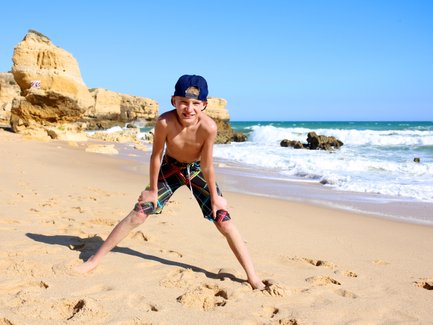 Ein fröhlicher Junge steht am Strand, mit weit gespreizten Beinen und einem breiten Lächeln, während die Wellen sanft an den Strand rollen.