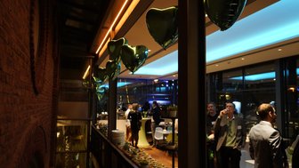 Im eleganten Raum hängen grüne Herzluftballons, während Gäste an einem Stehtisch miteinander plaudern und anstoßen.