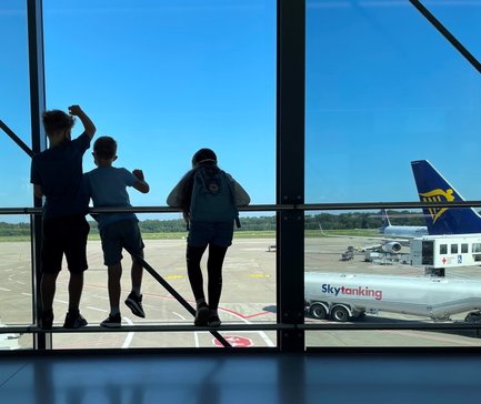 Drei Kinder stehen am Fenster eines Flughafens und beobachten das Geschehen auf dem Rollfeld bei strahlend blauem Himmel.