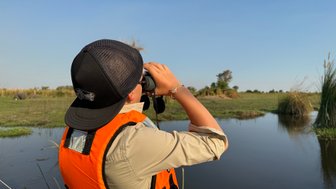 Ein Junge in einer orangefarbenen Weste beobachtet mit einem Fernglas die Landschaft am Wasser, umgeben von üppigem Grün.