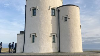 Ein historischer Turm mit runder Form und Zinnen steht vor einem klaren Himmel, während Besucher die Stufen hinaufgehen.