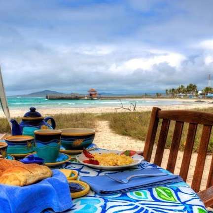 Ein gedeckter Tisch mit buntem Geschirr und frischem Brot steht am Strand, während die Wellen sanft im Hintergrund plätschern.