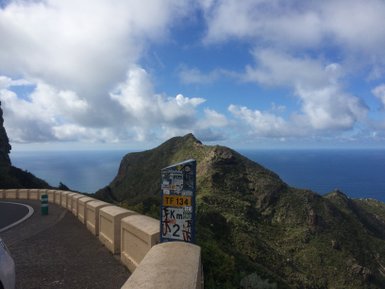 Eine kurvenreiche Straße führt entlang einer steilen Klippe mit Blick auf das glitzernde blaue Meer und bewaldete Hügel.