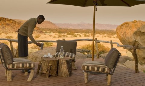Ein Mann gießt ein warmes Getränkt in eine Tasse auf der Terrasse des Camp Kipwe - Namibia Urlaub mit Kindern