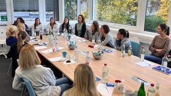 Eine Gruppe von Frauen sitzt an einem langen Tisch in einem hellen Raum, umgeben von großen Fenstern mit herbstlichen Bäumen.