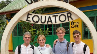 Eine Gruppe von vier Personen steht lächelnd am Äquator in Uganda, umgeben von bunten Gebäuden und Pflanzen.