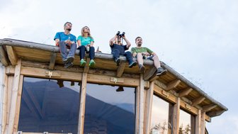 Vier Personen sitzen auf dem Dach einer Holzhütte und genießen die Aussicht, während sie Getränke in der Hand halten.