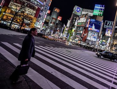 Ein Mann in Anzug überquert eine belebte Straße in Tokio, umgeben von leuchtenden Werbetafeln und nächtlicher Atmosphäre.