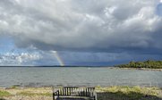 Eine einsame Bank steht am Ufer, während ein Regenbogen über dem ruhigen Wasser erscheint und die Wolken dramatisch wirken.